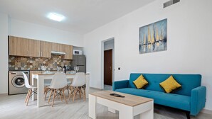 A modern kitchen with a dining area, a blue sofa with yellow cushions, and a painting of sailboats.