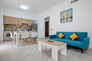 A modern kitchen with a dining area, a blue sofa with yellow cushions, and a painting of sailboats.