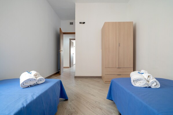 Une chambre avec deux lits, chacun recouvert d’une couverture bleue et agrémenté de serviettes blanches pliées. On y trouve une armoire en bois et une porte qui donne sur une autre pièce.
