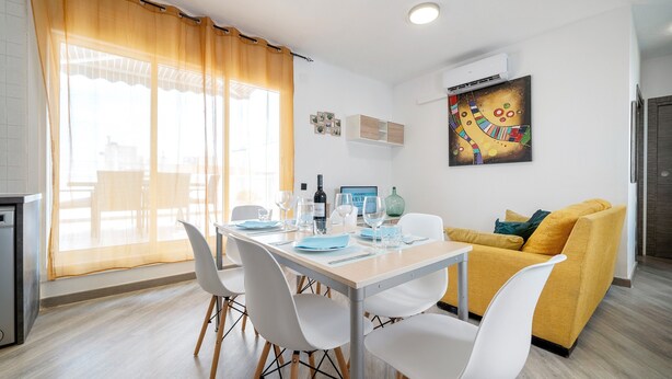 A modern living room with a dining area, a yellow sofa, and a white table set for four. - Holiday Apartment "Cambrils Turistic Atico" with Private Terrace & Wi-Fi (Cambrils)