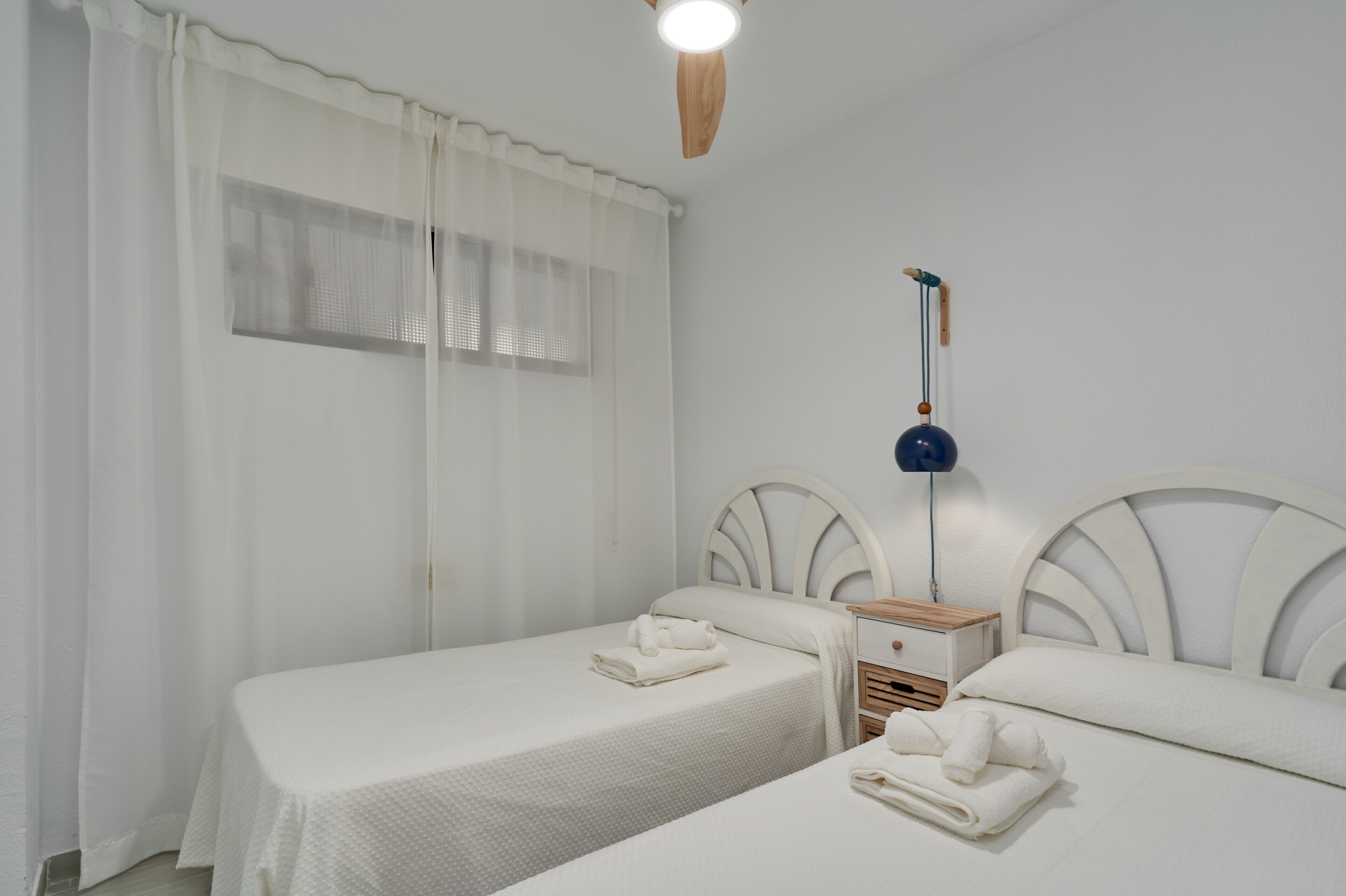 Una habitación con dos camas, una mesita de noche de madera, una lámpara de pared y una ventana con cortinas traslúcidas.
