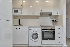 A compact kitchen with a microwave, oven, and washing machine, featuring white cabinetry and a tiled backsplash.