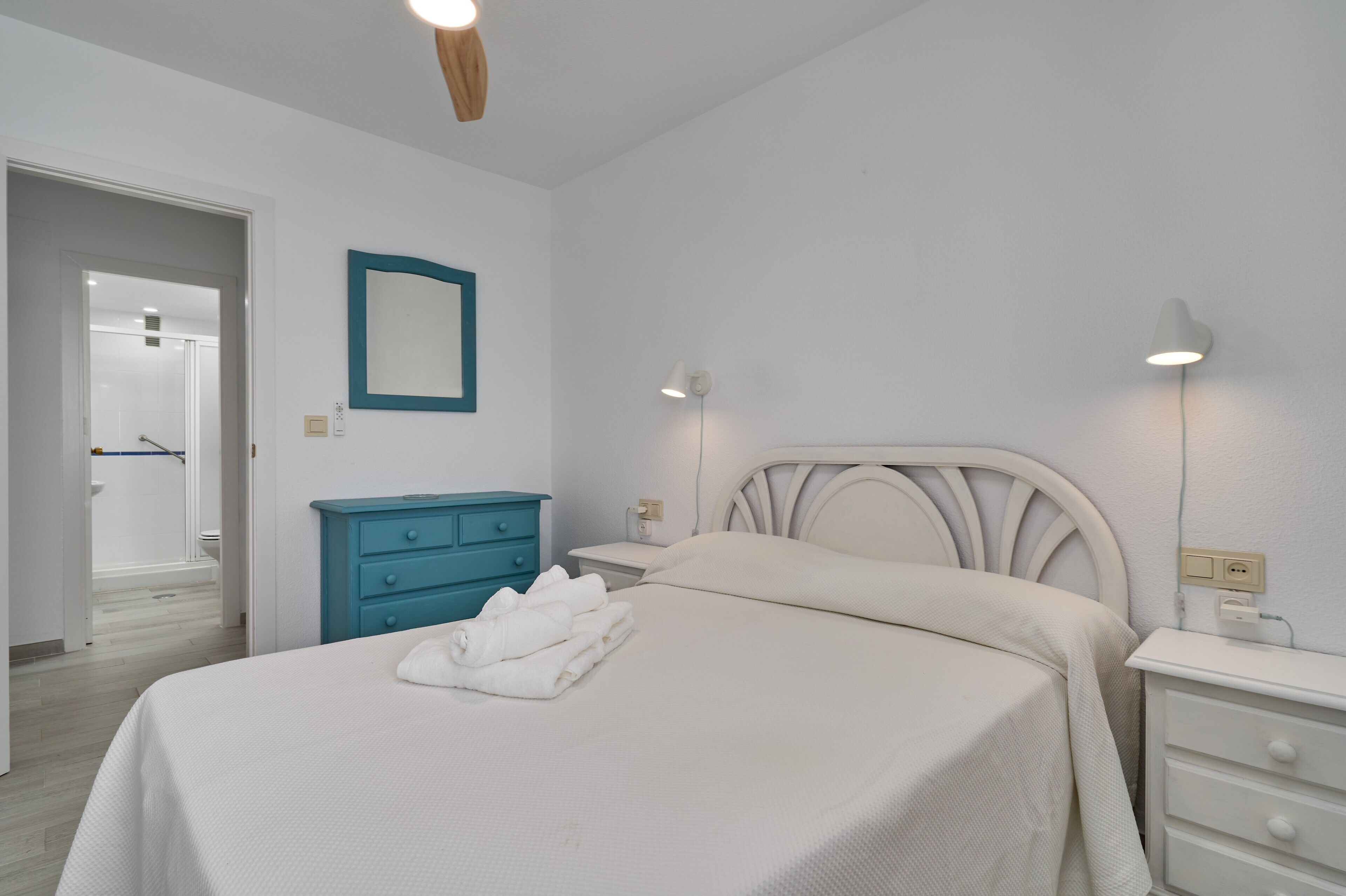 Un dormitorio con una cama, una mesita de noche, un aparador, un espejo y una puerta que da al baño.