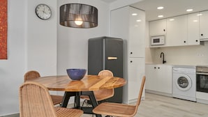 Moderni keittiö, jossa on ruokailutila. Tilaan kuuluu pyöreä puinen pöytä, punottu tuoli, musta jääkaappi, mikroaaltouuni ja pesukone.