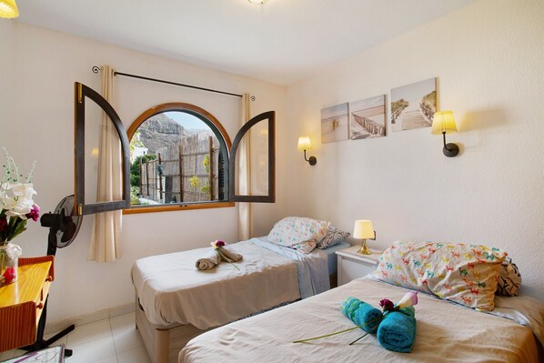 A hotel room with two beds, a window with curtains, a desk with a chair, and a wall-mounted lamp.