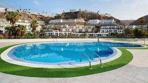A swimming pool with a surrounding green area, white buildings in the background, and a hilly landscape. - Vacation Apartment 'Residencial Sol Suite 2' with Sea View, Shared Pool and Wi-Fi (Puerto Rico)
