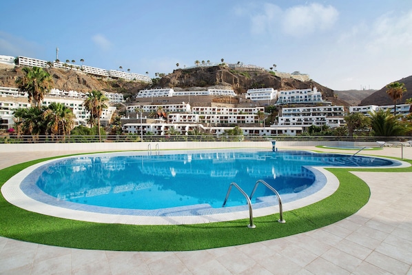A swimming pool with a surrounding green area, white buildings in the background, and a hilly landscape.