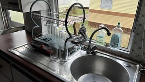 Una cocina con un lavamanos de acero inoxidable, un escurreplatos y una ventana con vista a una casa y una caravana.