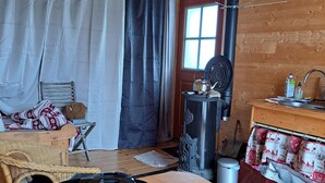 A cozy cabin interior with a wooden ceiling, a metal kettle on a table, a chair, a window with curtains, and a stove.