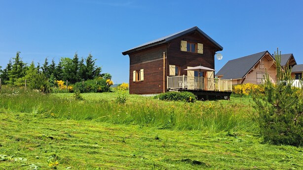 A wooden cabin with a balcony, surrounded by a grassy field and trees.