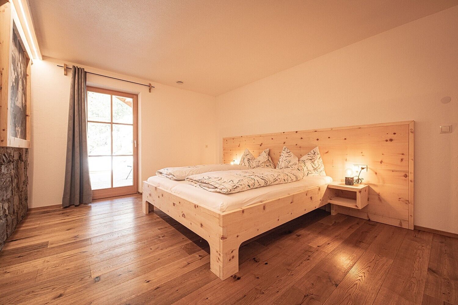 A bedroom with a wooden bed, a nightstand, a stone wall, and a large window with curtains.