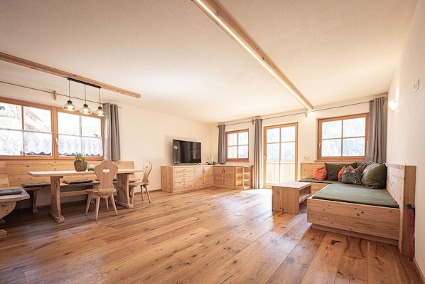 A spacious room with wooden flooring, a dining area with chairs and a table, a sofa with cushions, a television, and large windows.