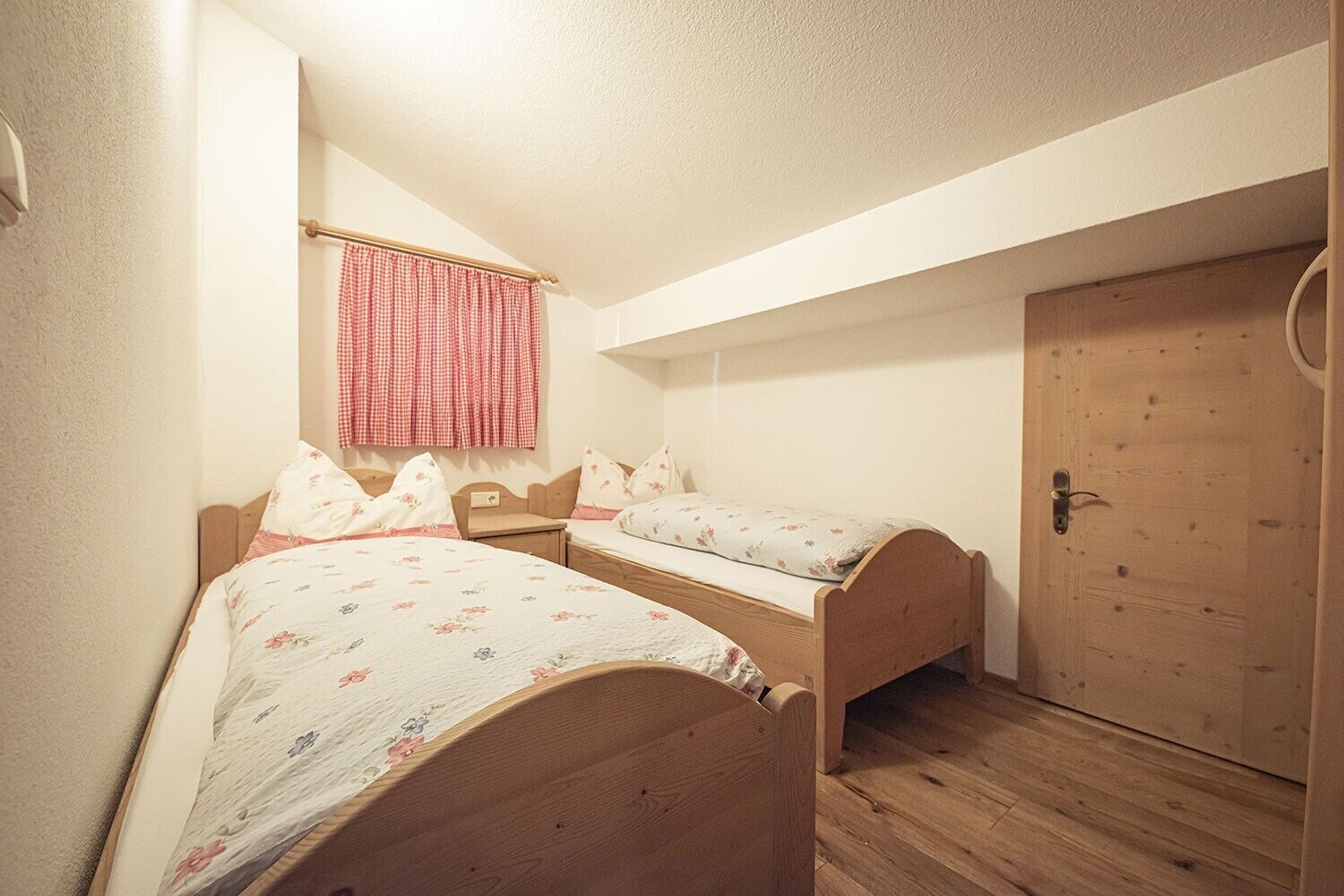 Un petit logement composé d’une seule pièce, avec deux lits en bois, une porte en bois et une fenêtre ornée d’un rideau à carreaux.