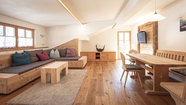 Een loft-achtige kamer met een houten bank, sofa en eettafel, houten vloeren en een aan de muur bevestigde televisie.