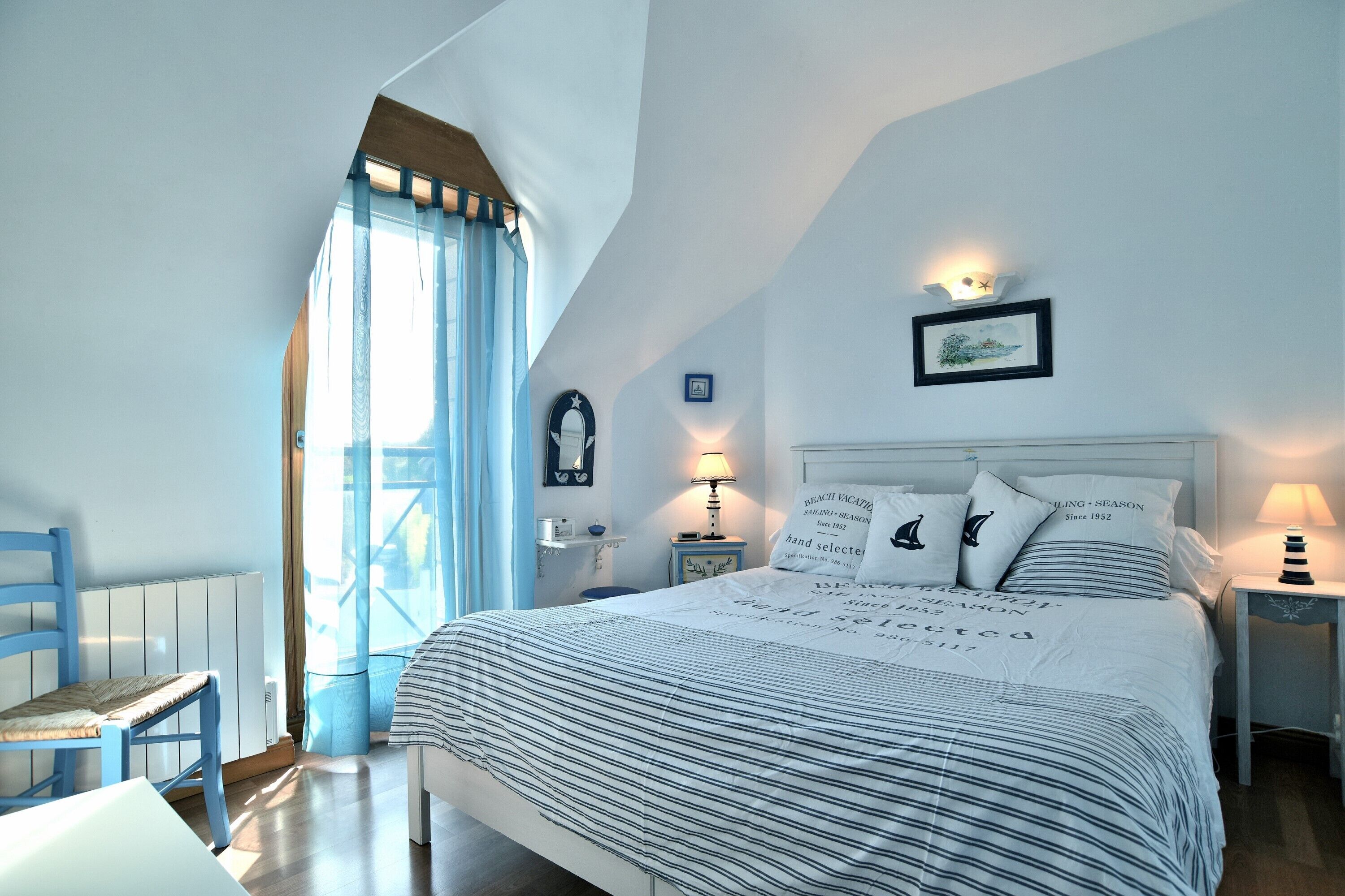 A bedroom with a bed, a chair, a nightstand, a lamp, a window with curtains, and a wall-mounted picture.