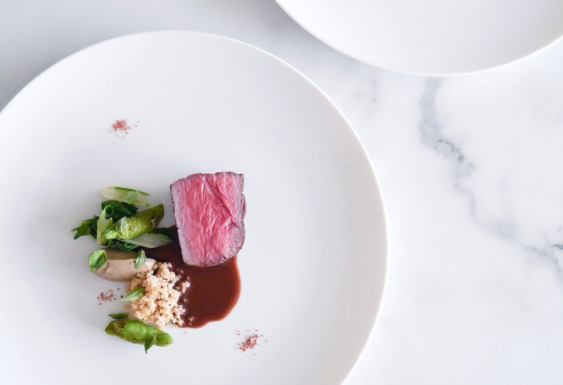 A white plate with a piece of meat, greens, and grains, accompanied by a small bowl of sauce.