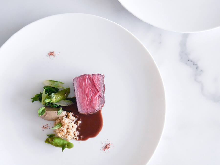 A white plate with a piece of meat, greens, and grains, accompanied by a small bowl of sauce.