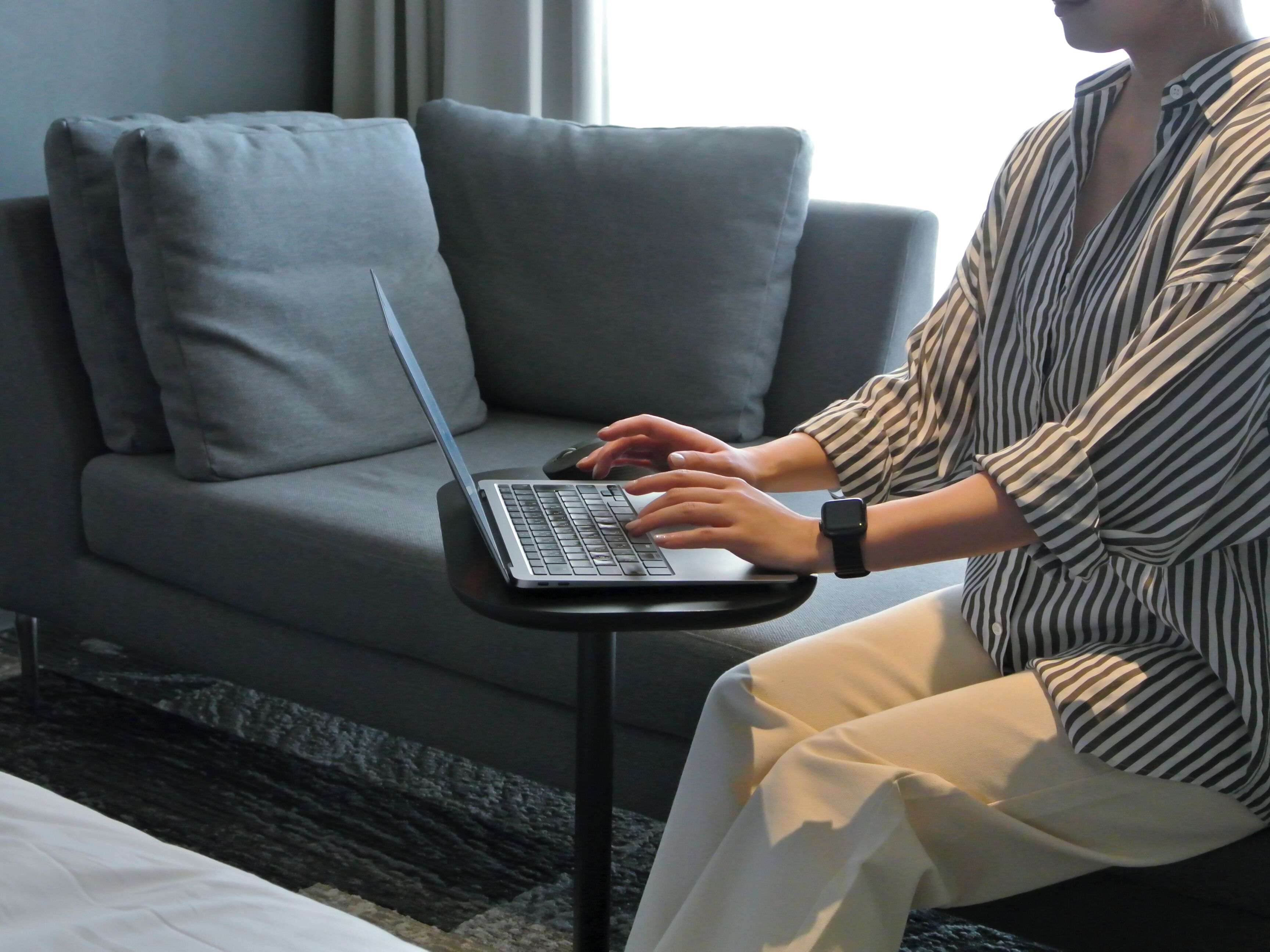 A person using a laptop on a small table in a room with a grey sofa and a window with curtains.