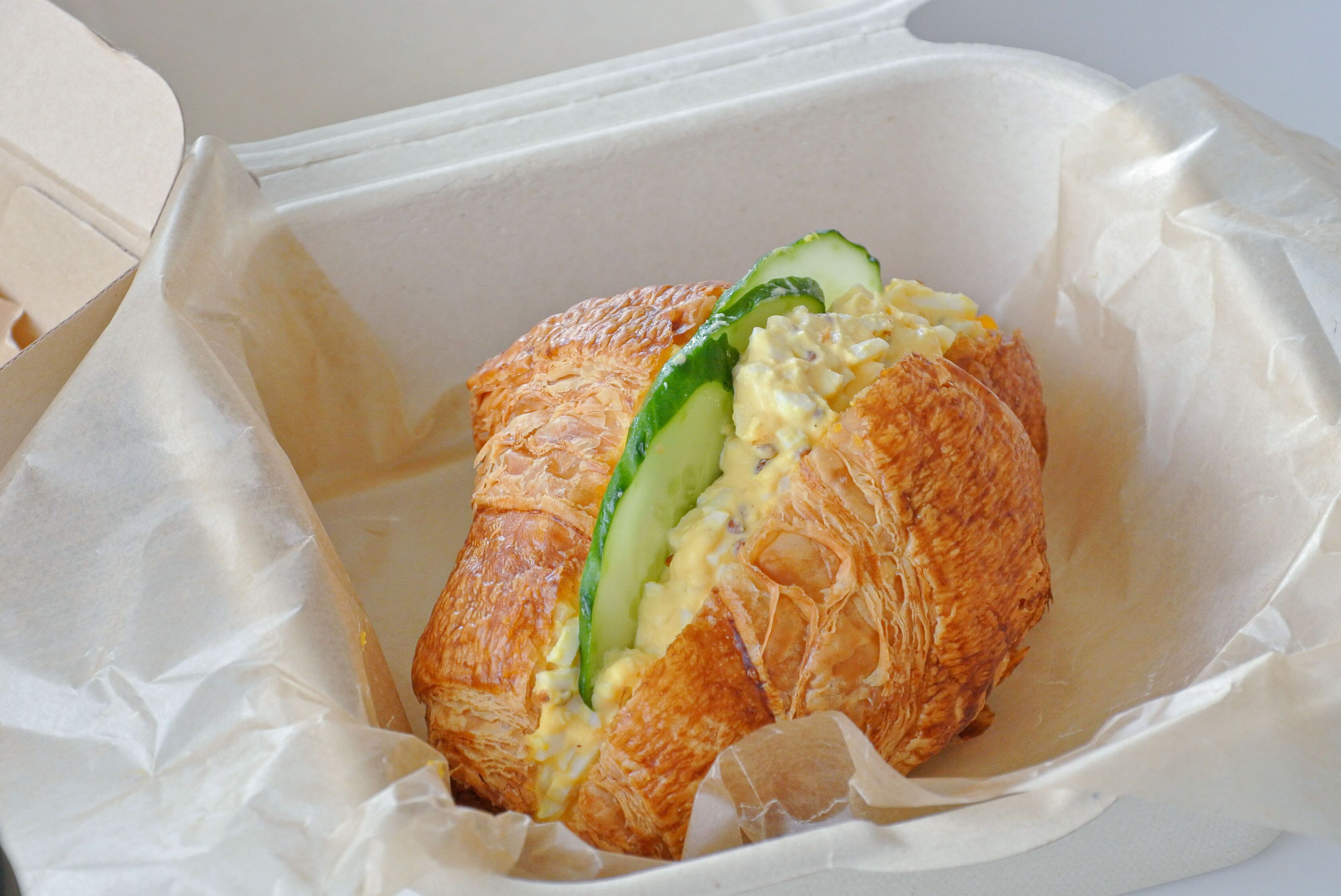 Un panino al croissant farcito con cetriolo a fette e crema, servito in un contenitore di carta bianca.