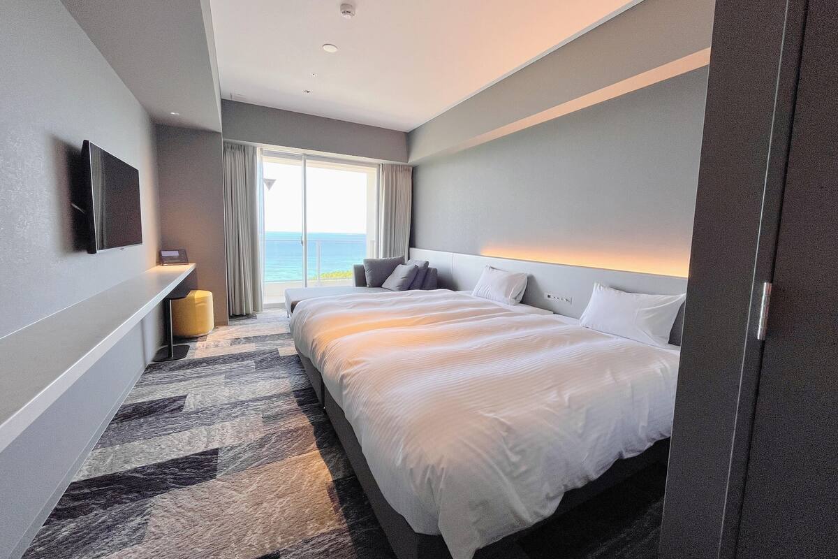 A modern hotel room with a large bed, a flat-screen TV, a bedside table, and a view of the ocean.