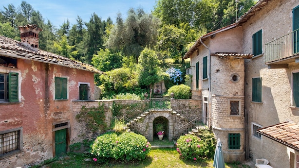 Um pátio com edifícios de pedra, um pequeno arco e vegetação exuberante.