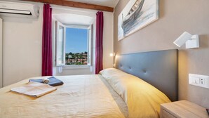 A hotel room with a bed, a nightstand, an air conditioning unit, a window with curtains, and a painting on the wall. - Holiday Apartment "Taronico Heron" with Lake View, Shared Terrace & Wi-Fi (bellagio)