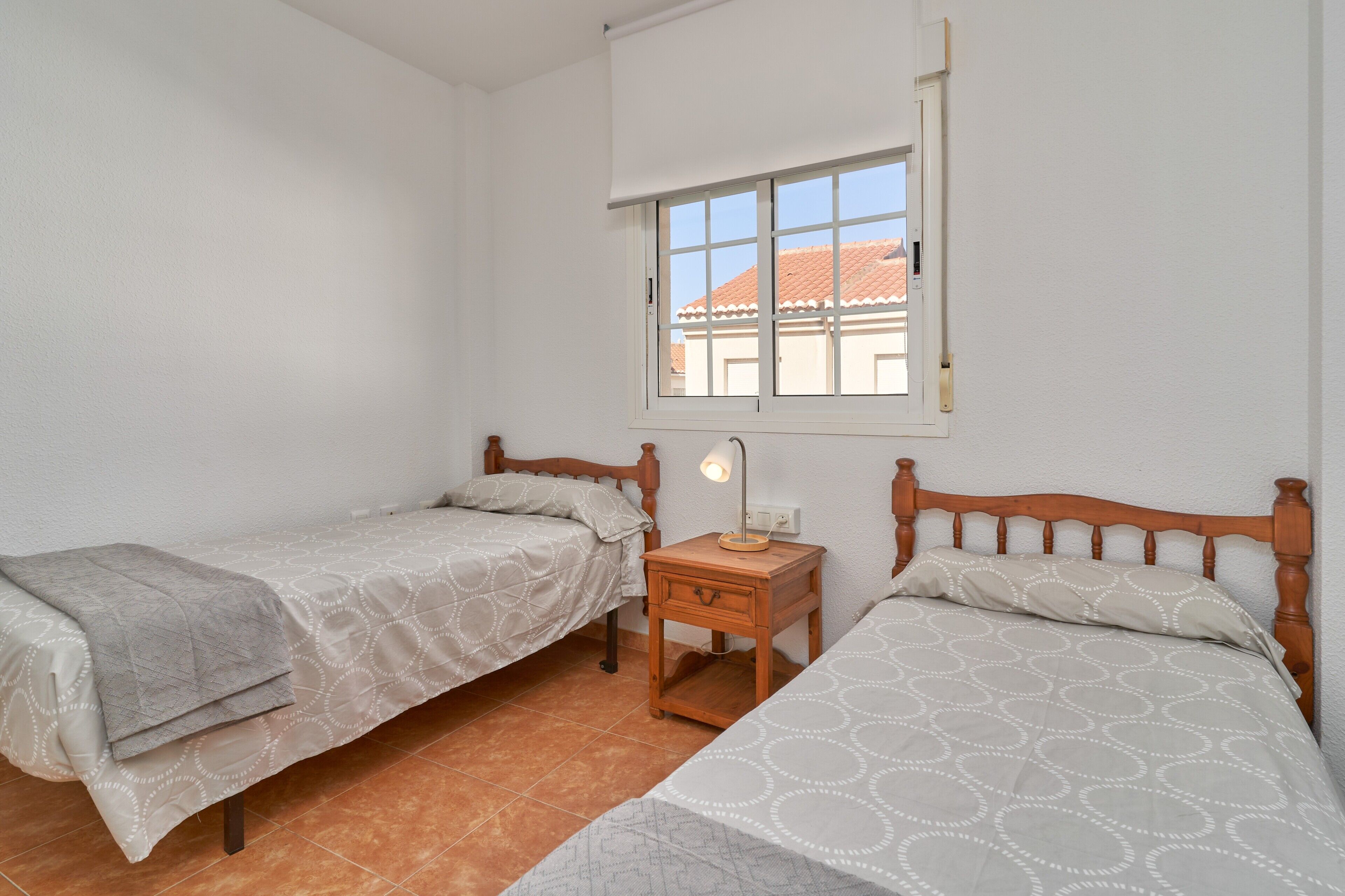 A bedroom with two single beds, a nightstand with a lamp, a window with a view of buildings, and a white wall.
