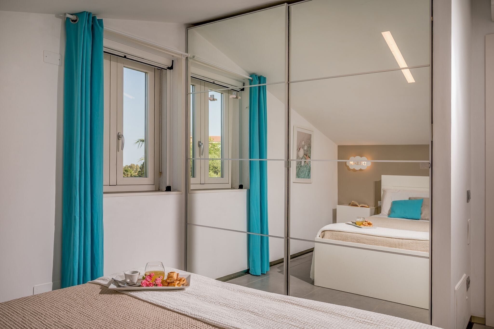 A bedroom with a bed, a bedside table with a tray of food, a window with curtains, and a mirror reflecting the room.