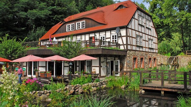 Un bâtiment traditionnel à colombages avec un toit rouge, une terrasse avec des parasols et un étang au premier plan.