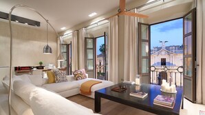 Un salon moderne avec un canapé, une table basse agrémentée de livres et de bougies, une lampe suspendue et une vue sur un bâtiment historique, visible à travers de grandes fenêtres.