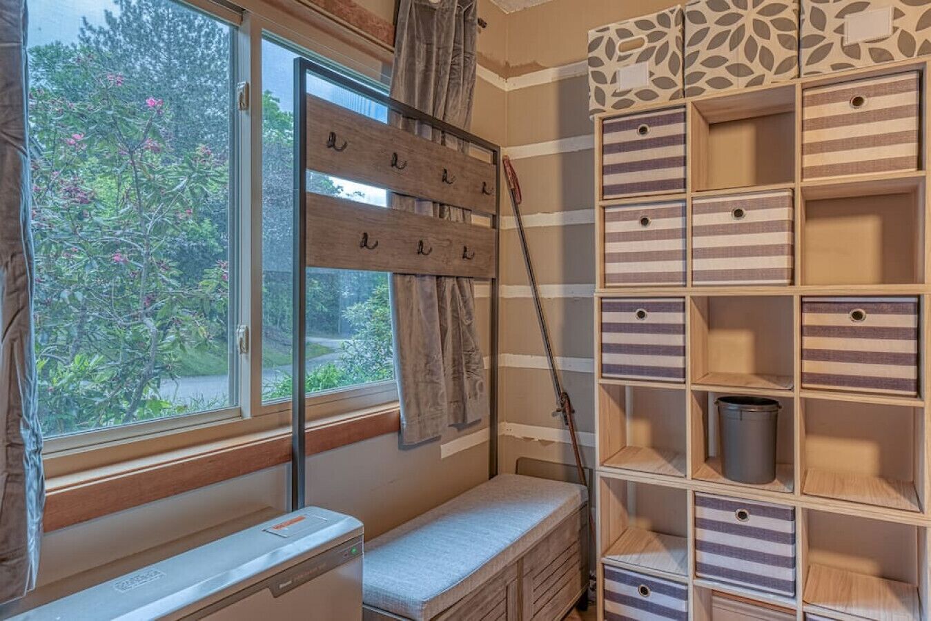 A room with a wooden desk, a bench, a window with curtains, and a storage unit with striped drawers.