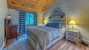 A bedroom with a wooden ceiling, a bed with a blue headboard, two bedside tables with lamps, a wooden dresser, and a window with patterned curtains.