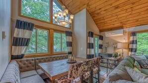 A cozy cabin interior with a wooden ceiling, large windows with striped curtains, a dining table, and a sofa.