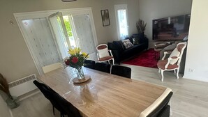 Un coin repas avec une table en bois, des chaises noires et un vase de fleurs, ainsi qu’un salon avec un canapé et un téléviseur en arrière-plan.