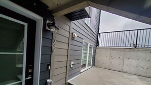 Un espacio exterior techado con suelo de hormigón, una puerta con panel de vidrio y una barandilla de metal.