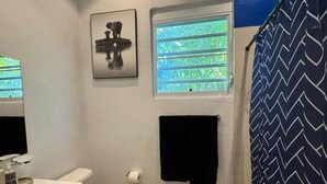 A bathroom with a white sink, a toilet, a window with a view of greenery, a black towel hanging on the wall, and a framed picture of a surfboard.