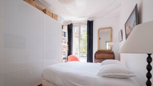 Une chambre à coucher comprenant un lit, une commode en bois, une table de chevet, une lampe, un miroir et une bibliothèque.