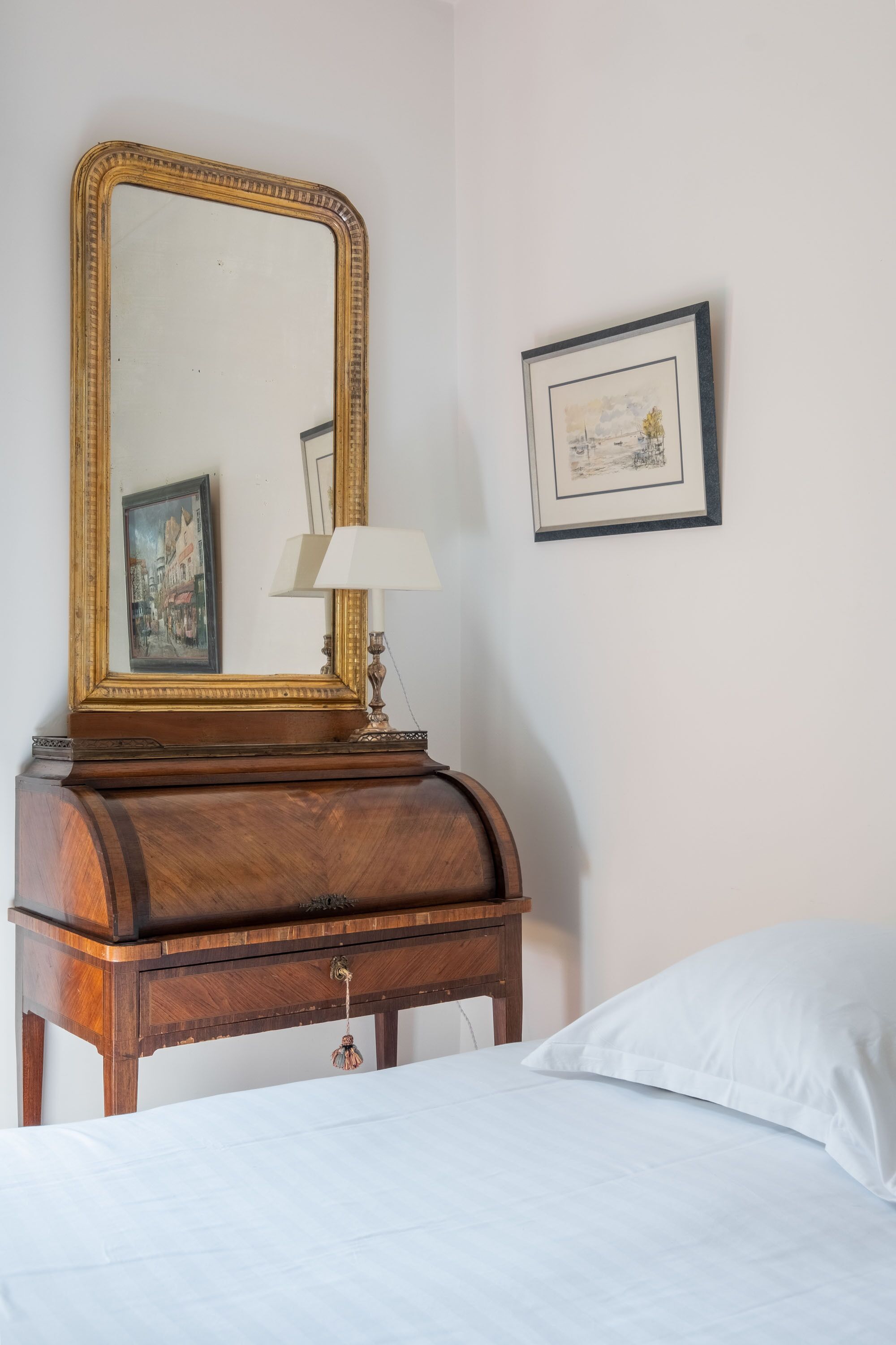A bedroom with a wooden dresser, a large mirror, a framed picture, a bedside lamp, and a white bed.