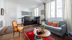 Un salon avec un canapé, une table basse et un fauteuil. Au fond, on aperçoit une cuisine avec un réfrigérateur et des placards.