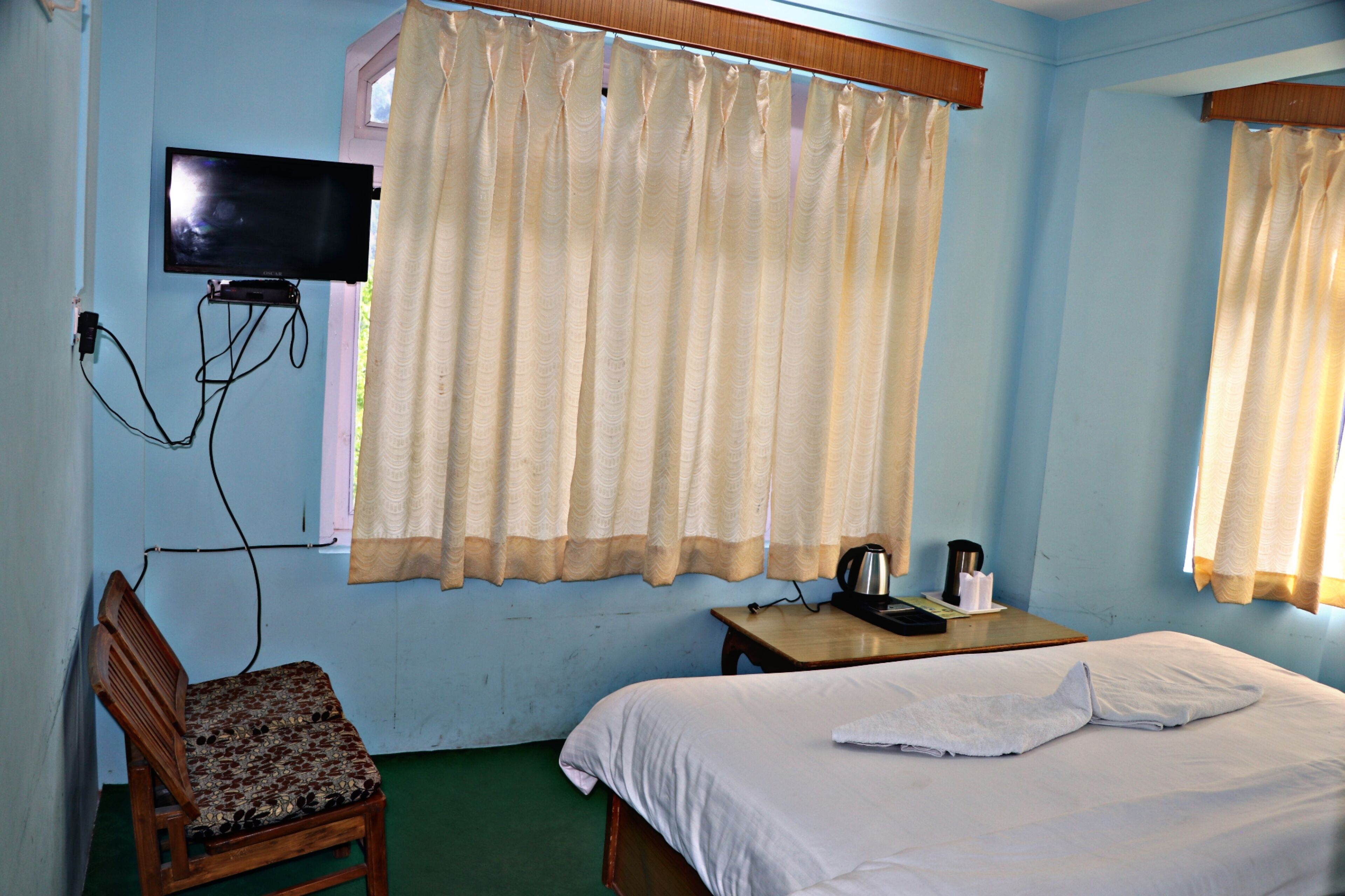 Une chambre d’hôtel avec un lit, une chaise, un téléviseur fixé au mur et une fenêtre avec des rideaux.