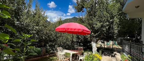 A patio with a red umbrella, a table, and chairs, surrounded by lush greenery and a clear blue sky.