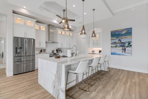 A modern kitchen with white cabinetry, a stainless steel refrigerator, a marble island with bar stools, and pendant lighting. - 706 NW 37th PL (Cape Coral)