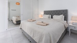 Une chambre moderne avec une tête de lit rembourrée de couleur grise, un grand lit avec une couette beige, deux lampes de chevet et un grand miroir.