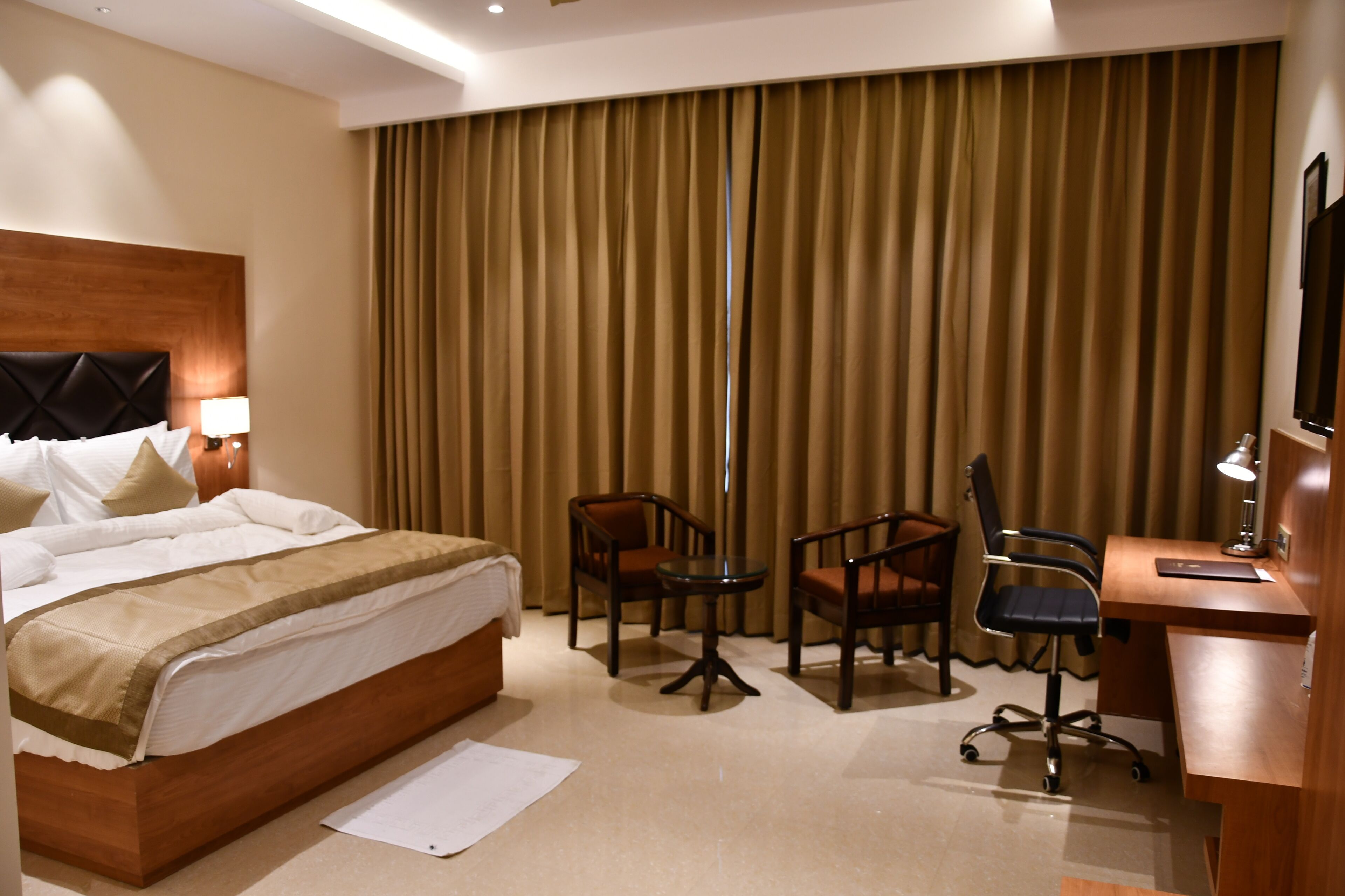 A hotel room with a bed, a desk, two chairs, a lamp, and a large window with curtains.