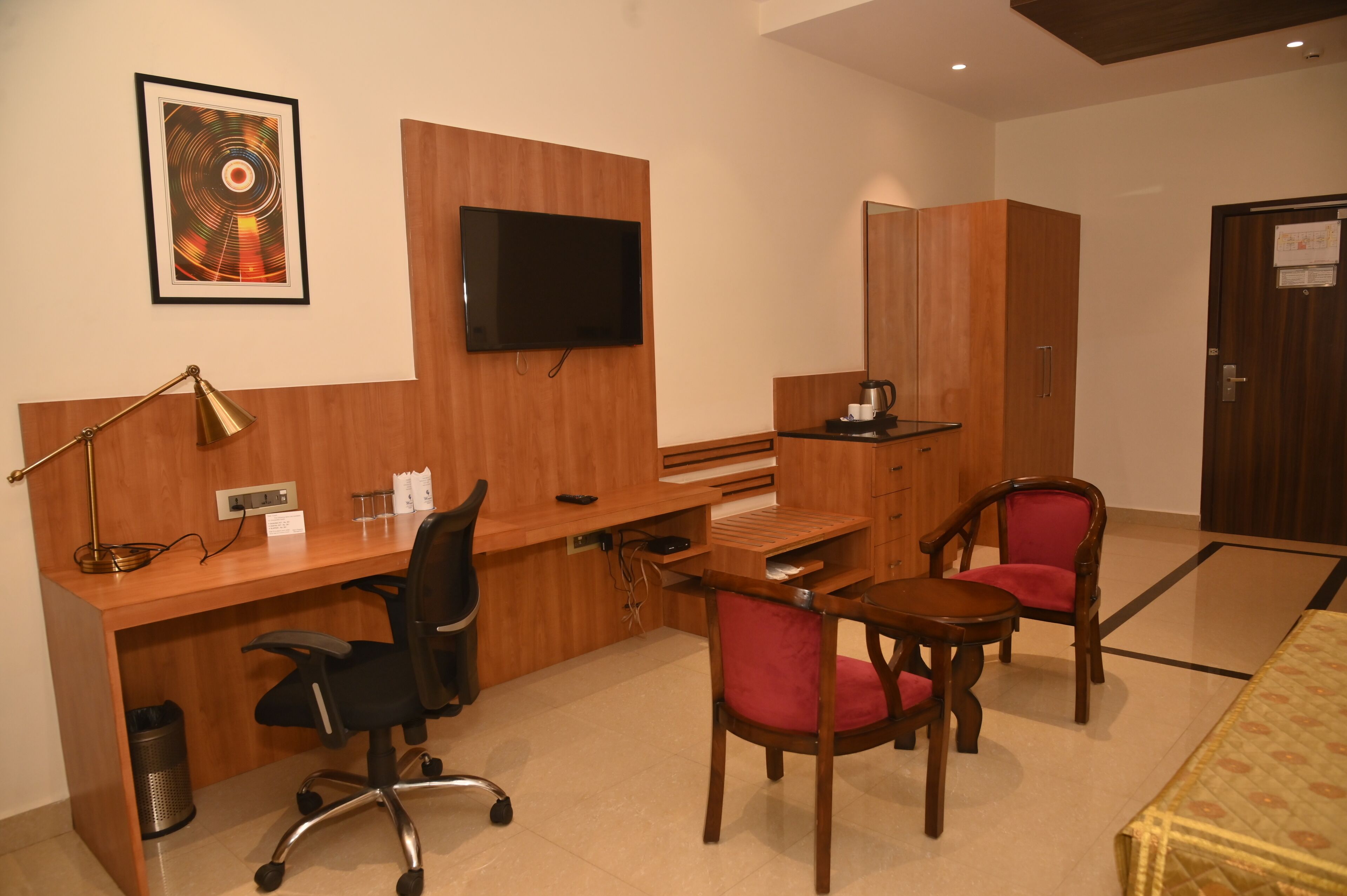 Une chambre d’hôtel avec un bureau en bois, un fauteuil de bureau noir, une chaise rouge, un téléviseur fixé au mur et une armoire en bois.