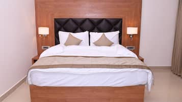 A hotel room with a wooden headboard, two bedside lamps, a bed with white bedding and two patterned pillows, and a small bedside table.