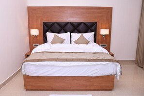 A hotel room with a wooden headboard, two bedside lamps, a bed with white bedding and two patterned pillows, and a small bedside table.