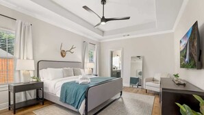 A bedroom with a bed, bedside tables, a ceiling fan, a window with curtains, a mirror, and a plant. - Poolside Place - Game Room - MTB-Friendly (Bentonville)