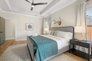Un dormitorio con cama, mesita de noche, ventilador de techo, un adorno de cabeza de venado en la pared y una ventana con cortinas.