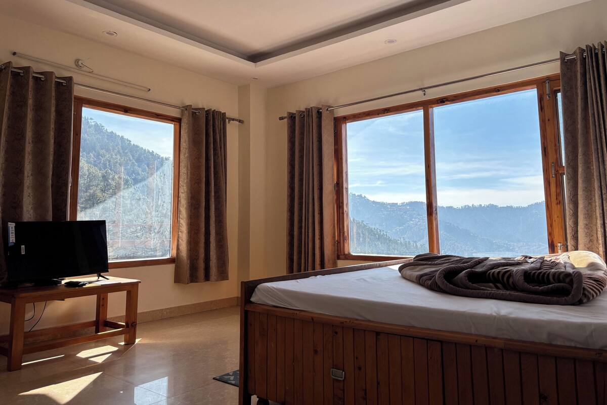 Un dormitorio con cama de madera, televisor sobre una mesa de madera y ventanas amplias con vistas a la montaña.